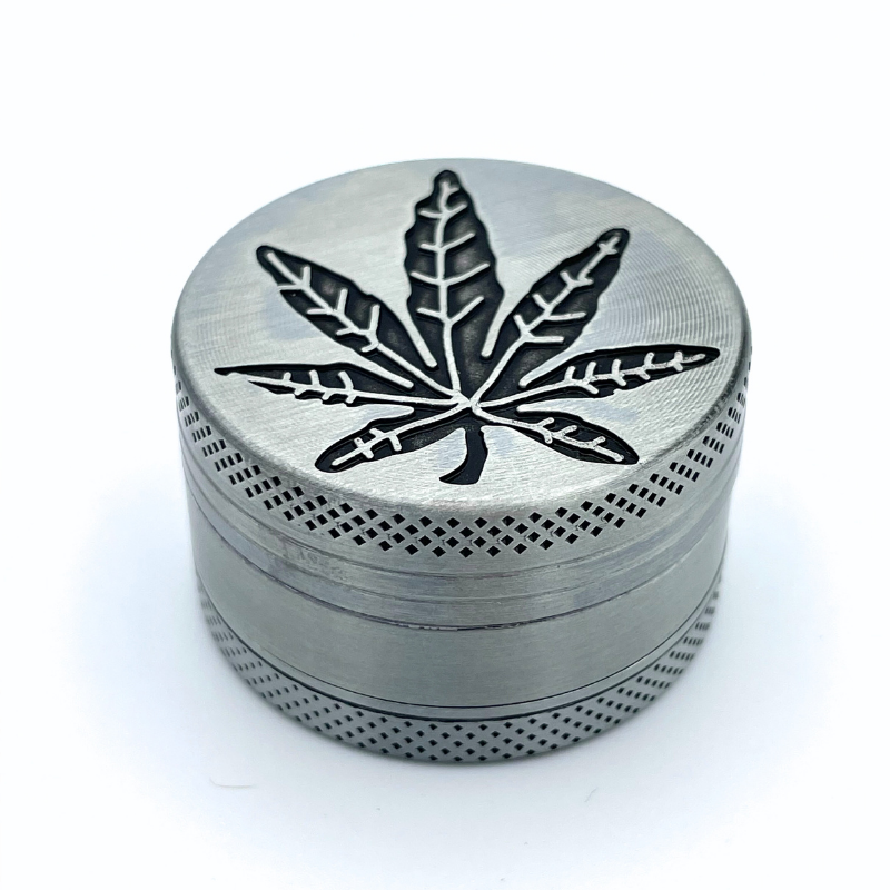 GRINDER METAL 3 PARTIES avec motif feuille de cannabis gravé sur le couvercle, idéal pour broyer les herbes. Accessoire robuste et élégant.